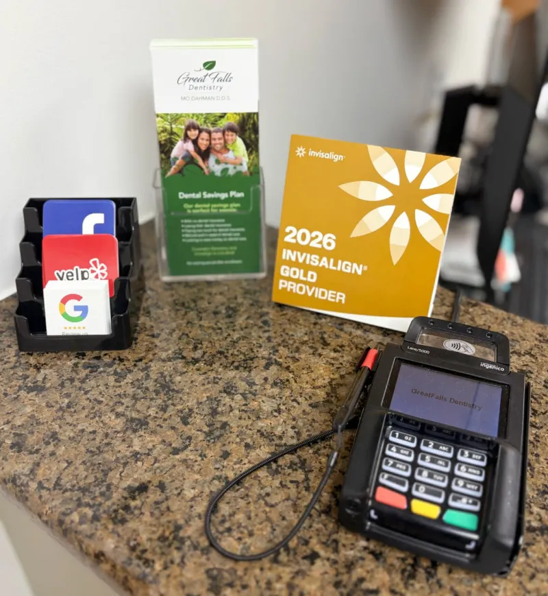 Dental office front desk counter with review cards for Facebook, Yelp, and Google, a Great Falls Dentistry brochure for their Dental Savings Plan, a 2026 Invisalign Gold Provider display card, and an Ingenico Lane/5000 payment terminal